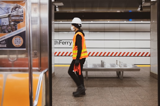 A person wearing a construction helmet, face mask, and high-visibility safety vest is walking on a subway platform. The background shows a tiled wall with red and white diagonal stripes and the word 'Ferry' partially visible. There is an empty bench and part of a subway car with an advertisement for the Brooklyn Half Marathon.
