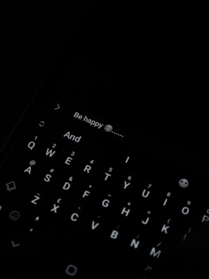 A dark image with a smartphone keyboard visible, displaying a message with the text 'Be happy 😊'. The keyboard is illuminated, showing a QWERTY layout.