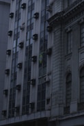 A building facade featuring a grid of windows with multiple air conditioning units installed on them. The structure has a modern design on the left side and a more classical, ornate style to the right. A flag with a distinctive green, yellow, and blue pattern is visible, indicating the country of origin.