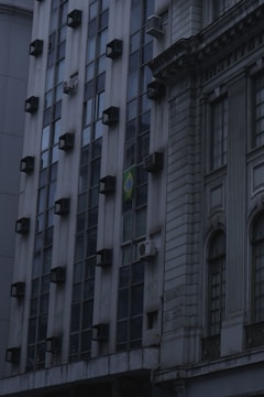 A building facade featuring a grid of windows with multiple air conditioning units installed on them. The structure has a modern design on the left side and a more classical, ornate style to the right. A flag with a distinctive green, yellow, and blue pattern is visible, indicating the country of origin.