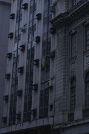 A building facade featuring a grid of windows with multiple air conditioning units installed on them. The structure has a modern design on the left side and a more classical, ornate style to the right. A flag with a distinctive green, yellow, and blue pattern is visible, indicating the country of origin.