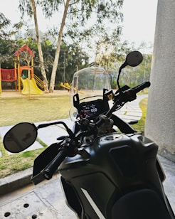 A motorcycle is parked near a playground. The handlebars and dashboard of the motorcycle are in the foreground, while in the background there is playground equipment including a slide and swings. The setting seems to be near a park area with grass and trees visible.