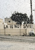 Raindrops are scattered across a window, with the blurred background showing a building and a message on a wall reading 'LA IS NOT SAFE'.