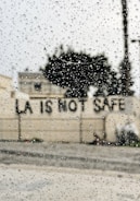 Raindrops are scattered across a window, with the blurred background showing a building and a message on a wall reading 'LA IS NOT SAFE'.