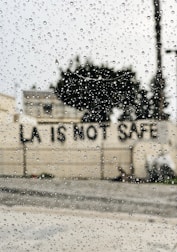Raindrops are scattered across a window, with the blurred background showing a building and a message on a wall reading 'LA IS NOT SAFE'.