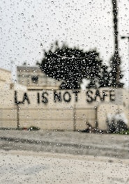 Raindrops are scattered across a window, with the blurred background showing a building and a message on a wall reading 'LA IS NOT SAFE'.