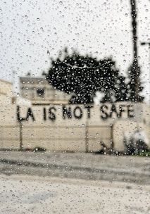 Raindrops are scattered across a window, with the blurred background showing a building and a message on a wall reading 'LA IS NOT SAFE'.
