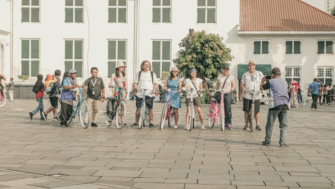 A group of people standing with bicycles in front of a historical building. They are posing for a photo, and a man is taking their picture. The building has white walls with green window shutters and a red tiled roof. Other people are seen in the background walking around the square.