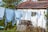 A row of assorted clothes hanging on a clothesline in a rural setting. The clothes are primarily light colors, set against a whitewashed wall of a small building with a red-tiled roof. The background includes lush greenery and a wooden stack. The atmosphere feels serene and rustic.