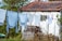 A row of assorted clothes hanging on a clothesline in a rural setting. The clothes are primarily light colors, set against a whitewashed wall of a small building with a red-tiled roof. The background includes lush greenery and a wooden stack. The atmosphere feels serene and rustic.