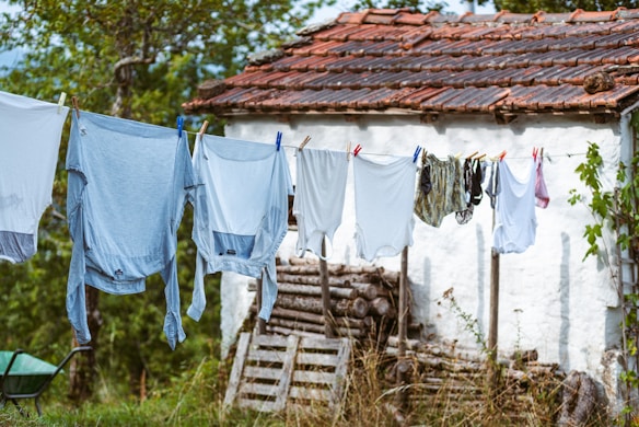 A row of assorted clothes hanging on a clothesline in a rural setting. The clothes are primarily light colors, set against a whitewashed wall of a small building with a red-tiled roof. The background includes lush greenery and a wooden stack. The atmosphere feels serene and rustic.