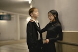 A young man and woman stand close together in a hallway. The man is wearing a black suit with a white shirt, and the woman is in a black dress. They seem to be engaged in a relaxed conversation, with the woman playfully holding the man's tie.