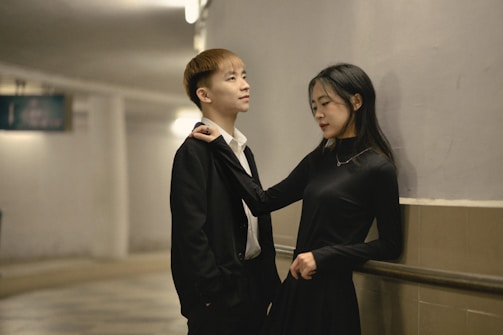 A young man and woman stand close together in a hallway. The man is wearing a black suit with a white shirt, and the woman is in a black dress. They seem to be engaged in a relaxed conversation, with the woman playfully holding the man's tie.