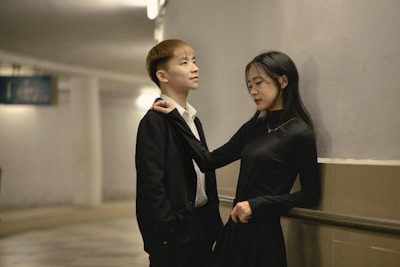 A young man and woman stand close together in a hallway. The man is wearing a black suit with a white shirt, and the woman is in a black dress. They seem to be engaged in a relaxed conversation, with the woman playfully holding the man's tie.