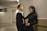 A young man and woman stand close together in a hallway. The man is wearing a black suit with a white shirt, and the woman is in a black dress. They seem to be engaged in a relaxed conversation, with the woman playfully holding the man's tie.