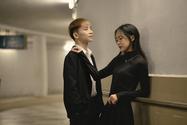 A young man and woman stand close together in a hallway. The man is wearing a black suit with a white shirt, and the woman is in a black dress. They seem to be engaged in a relaxed conversation, with the woman playfully holding the man's tie.