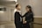 A young man and woman stand close together in a hallway. The man is wearing a black suit with a white shirt, and the woman is in a black dress. They seem to be engaged in a relaxed conversation, with the woman playfully holding the man's tie.