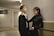 A young man and woman stand close together in a hallway. The man is wearing a black suit with a white shirt, and the woman is in a black dress. They seem to be engaged in a relaxed conversation, with the woman playfully holding the man's tie.