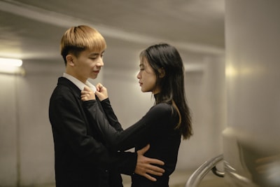A couple stands close together in an intimate setting, with soft lighting creating a warm atmosphere. The man has his arms around the woman, and they both appear to be engaged in a tender conversation.