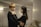 A couple stands close together in an intimate setting, with soft lighting creating a warm atmosphere. The man has his arms around the woman, and they both appear to be engaged in a tender conversation.