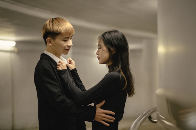 A couple stands close together in an intimate setting, with soft lighting creating a warm atmosphere. The man has his arms around the woman, and they both appear to be engaged in a tender conversation.