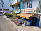 A street scene featuring multiple small shops with colorful signage, including a Vietnamese bakery or snack shop. The facade is adorned with striped awnings and potted plants. A narrow road passes by the shops. Decorative elements, like flowers and small trees, enhance the cozy atmosphere.