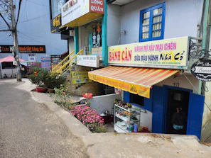 A street scene featuring multiple small shops with colorful signage, including a Vietnamese bakery or snack shop. The facade is adorned with striped awnings and potted plants. A narrow road passes by the shops. Decorative elements, like flowers and small trees, enhance the cozy atmosphere.