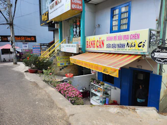 A street scene featuring multiple small shops with colorful signage, including a Vietnamese bakery or snack shop. The facade is adorned with striped awnings and potted plants. A narrow road passes by the shops. Decorative elements, like flowers and small trees, enhance the cozy atmosphere.