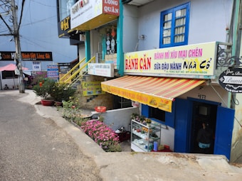 A street scene featuring multiple small shops with colorful signage, including a Vietnamese bakery or snack shop. The facade is adorned with striped awnings and potted plants. A narrow road passes by the shops. Decorative elements, like flowers and small trees, enhance the cozy atmosphere.