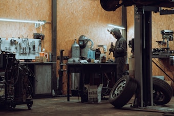 A workshop featuring a man in a hooded outfit working at a cluttered workbench. The space is equipped with various tools hanging on a pegboard, industrial equipment, tires, and a metal table. The lighting is subdued, with fluorescent fixtures providing illumination.