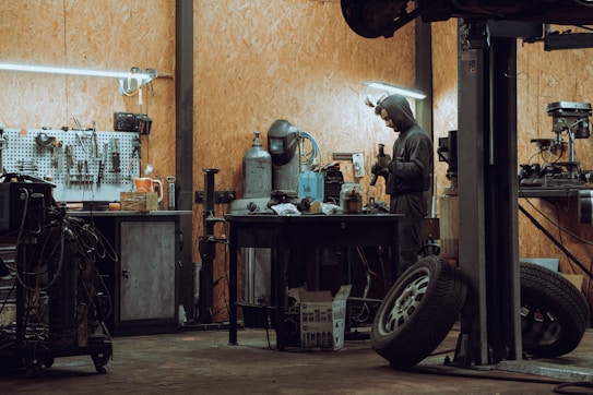A workshop featuring a man in a hooded outfit working at a cluttered workbench. The space is equipped with various tools hanging on a pegboard, industrial equipment, tires, and a metal table. The lighting is subdued, with fluorescent fixtures providing illumination.