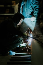 A person crouches while welding, creating bright sparks. Wearing a hooded jacket, the individual focuses intently on the task. An assistant stands nearby in work clothes, illuminated by the welding light in a dimly lit workshop.