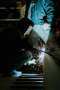 A person crouches while welding, creating bright sparks. Wearing a hooded jacket, the individual focuses intently on the task. An assistant stands nearby in work clothes, illuminated by the welding light in a dimly lit workshop.
