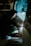 A person crouches while welding, creating bright sparks. Wearing a hooded jacket, the individual focuses intently on the task. An assistant stands nearby in work clothes, illuminated by the welding light in a dimly lit workshop.
