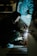 A person crouches while welding, creating bright sparks. Wearing a hooded jacket, the individual focuses intently on the task. An assistant stands nearby in work clothes, illuminated by the welding light in a dimly lit workshop.