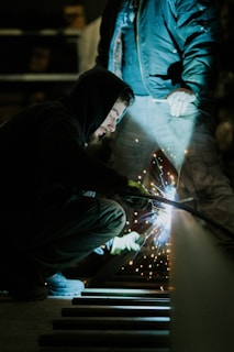 A person crouches while welding, creating bright sparks. Wearing a hooded jacket, the individual focuses intently on the task. An assistant stands nearby in work clothes, illuminated by the welding light in a dimly lit workshop.