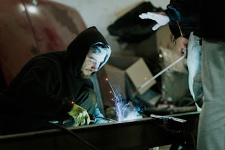A person in a dark hoodie is intently focused on welding, with a bright blue light and sparks emanating from the work area. Another person nearby is observing, partially visible with a hand extended. The setting appears to be a workshop with various metal objects and tools around.