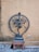 A bronze statue of a dancing deity, known as Nataraja, with multiple arms and one leg lifted, placed on a small stone platform. Encircling the figure is a decorative, flame-adorned aureole. The background features a plain, weathered wall and stone flooring.