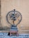 A bronze statue of a dancing deity, known as Nataraja, with multiple arms and one leg lifted, placed on a small stone platform. Encircling the figure is a decorative, flame-adorned aureole. The background features a plain, weathered wall and stone flooring.