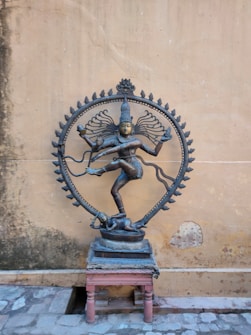 A bronze statue of a dancing deity, known as Nataraja, with multiple arms and one leg lifted, placed on a small stone platform. Encircling the figure is a decorative, flame-adorned aureole. The background features a plain, weathered wall and stone flooring.