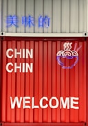 A brightly colored red and white metal wall featuring neon signage and text. The neon displays a stylized steaming bowl of noodles alongside Chinese characters and the phrases 'CHIN CHIN' and 'WELCOME' in bold letters.