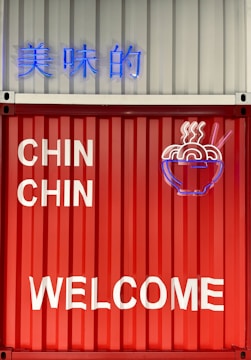 A brightly colored red and white metal wall featuring neon signage and text. The neon displays a stylized steaming bowl of noodles alongside Chinese characters and the phrases 'CHIN CHIN' and 'WELCOME' in bold letters.