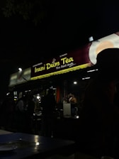 A dimly lit roadside tea stall in the evening featuring signage for 'Irani Dum Tea'. Several people are gathered around the stall, some possibly ordering or waiting for tea. The atmosphere is casual and busy with the presence of tables and chairs in the foreground.