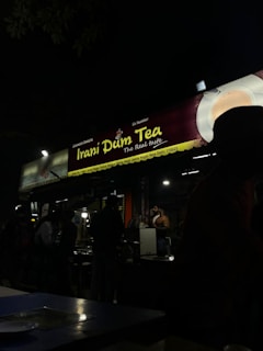 A dimly lit roadside tea stall in the evening featuring signage for 'Irani Dum Tea'. Several people are gathered around the stall, some possibly ordering or waiting for tea. The atmosphere is casual and busy with the presence of tables and chairs in the foreground.