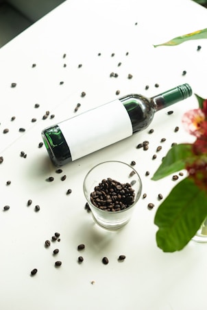 A wine bottle lies on a light surface, surrounded by scattered coffee beans. A glass filled with coffee beans is positioned in the foreground. To the right, a flower with red petals and green leaves partially enters the frame.