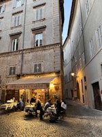 An outdoor restaurant setting with people dining at tables under a canopy labeled 'Osteria Del Gallo.' The scene is situated in a narrow, cobblestone alleyway between tall, old buildings with shuttered windows. Warm lighting creates a cozy and inviting atmosphere.