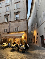 An outdoor restaurant setting with people dining at tables under a canopy labeled 'Osteria Del Gallo.' The scene is situated in a narrow, cobblestone alleyway between tall, old buildings with shuttered windows. Warm lighting creates a cozy and inviting atmosphere.