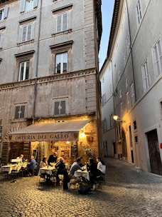 An outdoor restaurant setting with people dining at tables under a canopy labeled 'Osteria Del Gallo.' The scene is situated in a narrow, cobblestone alleyway between tall, old buildings with shuttered windows. Warm lighting creates a cozy and inviting atmosphere.