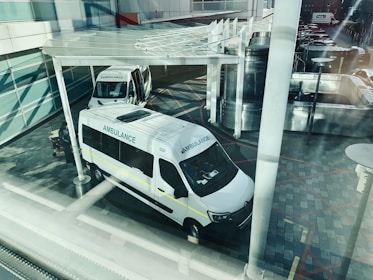 Two ambulances are parked outside what appears to be a modern building. The view is from above and through a window, creating reflections and a slightly obstructed view. The ground is paved with patterned tiles and there is a partially visible shelter above the ambulances. The scene appears calm with no immediate activity around the vehicles.