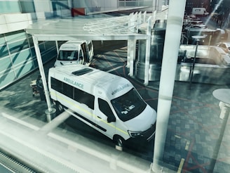 Two ambulances are parked outside what appears to be a modern building. The view is from above and through a window, creating reflections and a slightly obstructed view. The ground is paved with patterned tiles and there is a partially visible shelter above the ambulances. The scene appears calm with no immediate activity around the vehicles.